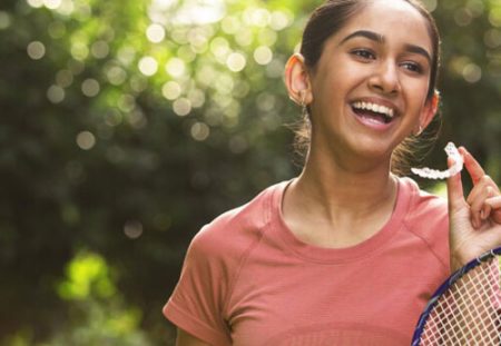 Hispanic teenage girl outdoors holding an Invisalign tray and a sports racket.